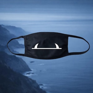 Op de afbeelding: Zwart gezichtsmasker met een wit grafisch ontwerp. Het ontwerp heeft een horizontale lijn met twee gebogen lijnen erboven. Het masker heeft zwarte oorlussen en staat tegen een vage achtergrond van bergen en water.