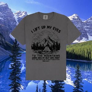 Könnte beinhalten: Ein graues T-Shirt mit einem Berg- und Zelt-Motiv. Der Text lautet: "Ich hebe meine Augen auf zu den Bergen, woher kommt mir Hilfe?" Psalm 121:1-2 ist ebenfalls aufgedruckt. Das T-Shirt hat einen Rundhalsausschnitt und kurze Ärmel.