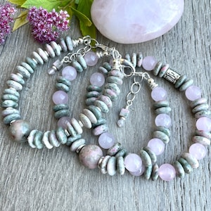 May include: Three bracelets made with green and pink gemstone beads and silver clasps. The bracelets are stacked on a wooden surface.