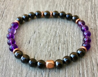 May include: A beaded bracelet featuring a repeating pattern of smooth, round black beads and translucent purple beads. The design is accented with small, metallic copper-colored spacer beads and a single, larger, square-shaped copper-colored bead.