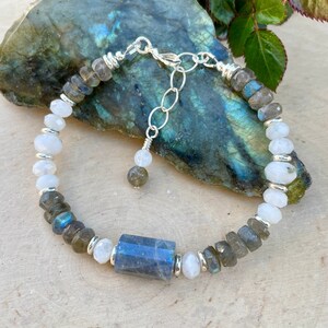 May include: A silver bracelet with white and gray gemstone beads. The bracelet has a larger, rectangular, blue-green gemstone in the center.