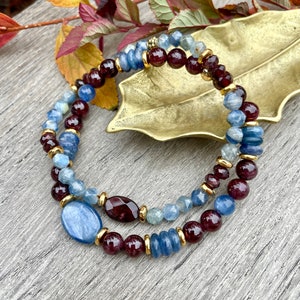 May include: Two beaded bracelets. One bracelet features blue and maroon beads with gold accents. The other bracelet has blue and maroon beads with a large oval blue stone and gold accents. The bracelets are on a wooden surface.