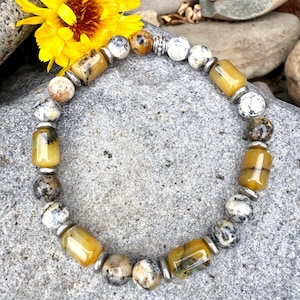 May include: A beaded bracelet featuring a mix of yellow, white, and speckled beads. The bracelet includes cylindrical and spherical beads, separated by silver-colored metal spacers. A bright yellow flower is visible in the background.