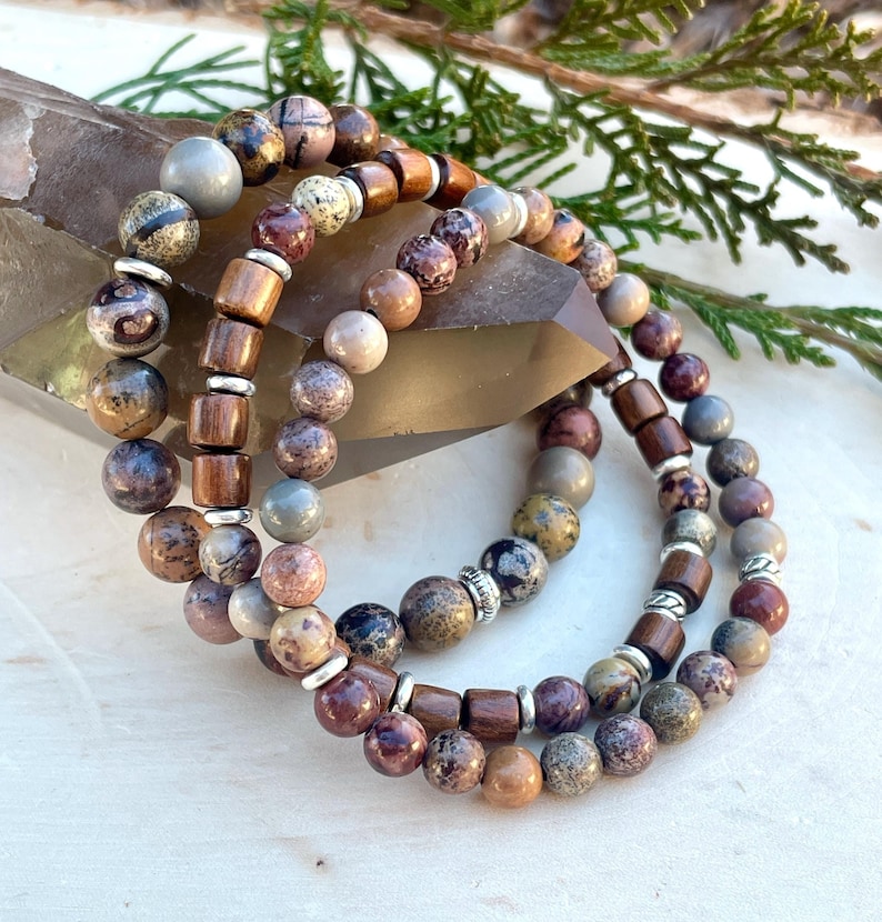 K&ouml;nnte beinhalten: Drei &uuml;bereinanderliegende Armb&auml;nder mit braunen, grauen und wei&szlig;en Steinperlen und silbernen Akzenten. Die Armb&auml;nder sind aus Holzperlen und Naturstein gefertigt.