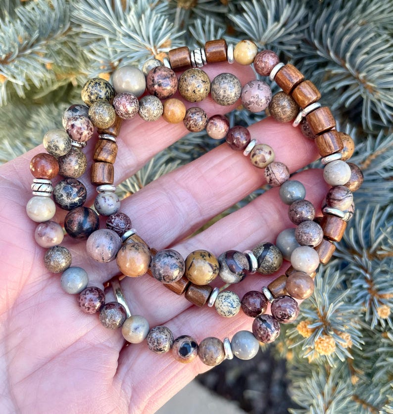 K&ouml;nnte beinhalten: Drei &uuml;bereinanderliegende Armb&auml;nder mit braunen, grauen und wei&szlig;en gesprenkelten Perlen. Die Perlen sind mit silbernen und braunen Abstandshaltern durchsetzt.