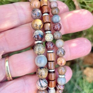 K&ouml;nnte beinhalten: Drei &uuml;bereinanderliegende Armb&auml;nder mit braunen Holzperlen, grauen und braunen gesprenkelten Perlen und silbernen Abstandshaltern.