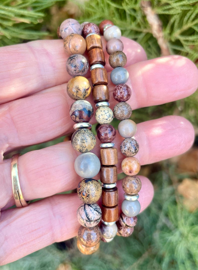 K&ouml;nnte beinhalten: Drei &uuml;bereinanderliegende Armb&auml;nder mit braunen Holzperlen, grauen und braunen gesprenkelten Perlen und silbernen Abstandshaltern.