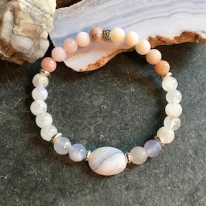 May include: A bracelet made with white, pink, and blue gemstone beads. The bracelet has a silver charm and a larger, oval-shaped blue agate bead.