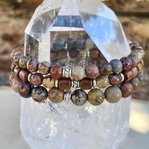 K&ouml;nnte beinhalten: Drei &uuml;bereinanderliegende Armb&auml;nder aus braunen, beigen und roten Jaspisperlen mit silbernen Zwischenst&uuml;cken. Die Armb&auml;nder werden auf einem klaren Quarzkristall pr&auml;sentiert.