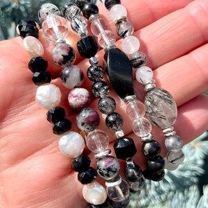 Peut inclure: Un ensemble de six bracelets avec des perles noires, blanches et grises. Les bracelets sont faits de diff&eacute;rents mat&eacute;riaux, dont l'onyx noir, les perles blanches et les cristaux de quartz transparents. Certaines des perles ont une finition scintillante.
