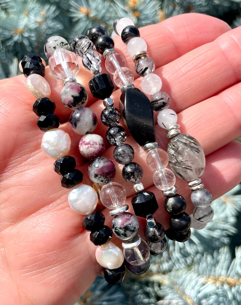 Peut inclure: Un ensemble de six bracelets avec des perles noires, blanches et grises. Les bracelets sont faits de diff&eacute;rents mat&eacute;riaux, dont l'onyx noir, les perles blanches et les cristaux de quartz transparents. Certaines des perles ont une finition scintillante.