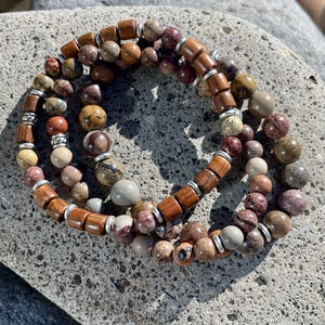 K&ouml;nnte beinhalten: Drei &uuml;bereinanderliegende Armb&auml;nder mit braunen Holzperlen und mehrfarbigen Steinperlen. Die Armb&auml;nder liegen auf einer grauen Steinoberfl&auml;che.