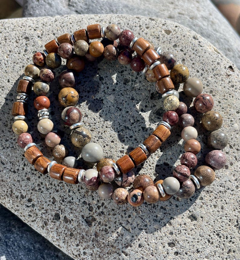 K&ouml;nnte beinhalten: Drei &uuml;bereinanderliegende Armb&auml;nder mit braunen Holzperlen und mehrfarbigen Steinperlen. Die Armb&auml;nder liegen auf einer grauen Steinoberfl&auml;che.