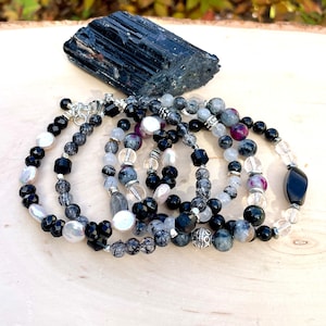 Peut inclure: Un ensemble de cinq bracelets en onyx noir, quartz clair et pierres pr&eacute;cieuses grises. Les bracelets sont empil&eacute;s sur une surface en bois avec un cristal de tourmaline noire en arri&egrave;re-plan.