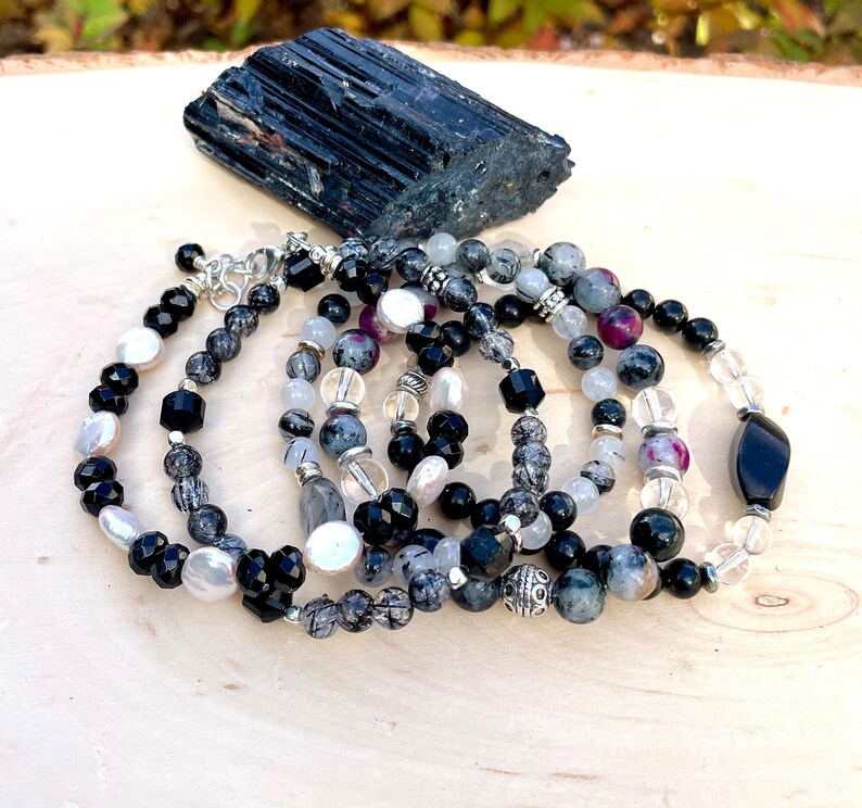 Peut inclure: Un ensemble de cinq bracelets en onyx noir, quartz clair et pierres pr&eacute;cieuses grises. Les bracelets sont empil&eacute;s sur une surface en bois avec un cristal de tourmaline noire en arri&egrave;re-plan.