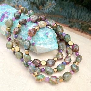 Pode incluir: Um colar de várias voltas feito com contas de pedras preciosas verdes, marrons e rosadas e espaçadores dourados. O colar é apresentado em uma ponta de cristal verde e branca.