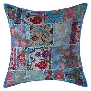 May include: Square decorative pillow with a patchwork design. The pillow features a light blue background with various squares of fabric in shades of blue, red, pink, and gold. Floral and paisley patterns adorn the squares, with a solid blue border.
