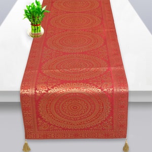 May include: Red table runner with a gold mandala pattern. The runner has a fringed edge and tassels on the ends.