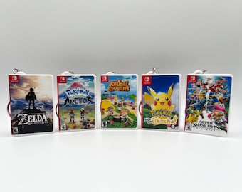 Pode incluir: Cinco chaveiros de jogos diferentes do Nintendo Switch. Os chaveiros são brancos com imagens coloridas dos jogos. Os jogos são The Legend of Zelda: Breath of the Wild, Pokemon Legends: Arceus, Animal Crossing: New Horizons, Pokemon Let's Go Pikachu e Super Smash Bros. Ultimate.
