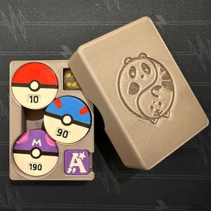 Pode incluir: Uma caixa de armazenamento cinza impressa em 3D com um design de panda yin-yang na tampa. Dentro h&aacute; tr&ecirc;s fichas circulares que lembram Pok&eacute; Balls com os n&uacute;meros 10, 90 e 190, uma ficha quadrada roxa com a letra A e um dado amarelo transl&uacute;cido.