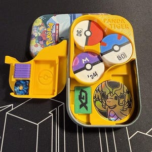 Pode incluir: Um inserto de pl&aacute;stico amarelo dentro de uma lata de metal para um jogo de cartas colecion&aacute;veis Pokemon. O inserto cont&eacute;m tr&ecirc;s fichas circulares com os n&uacute;meros 30, 80 e 24, um marcador GX retangular verde, um dado azul, cubos roxos e uma carta de drag&atilde;o hologr&aacute;fica.