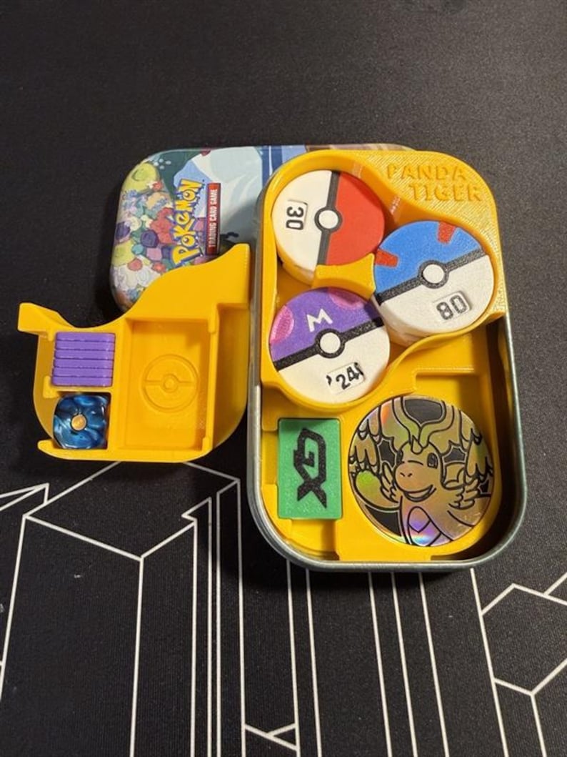 Pode incluir: Um inserto de pl&aacute;stico amarelo dentro de uma lata de metal para um jogo de cartas colecion&aacute;veis Pokemon. O inserto cont&eacute;m tr&ecirc;s fichas circulares com os n&uacute;meros 30, 80 e 24, um marcador GX retangular verde, um dado azul, cubos roxos e uma carta de drag&atilde;o hologr&aacute;fica.