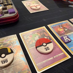 Pode incluir: Um conjunto de acess&oacute;rios para jogos de cartas colecion&aacute;veis, incluindo tr&ecirc;s contadores de dano circulares de pl&aacute;stico com detalhes em vermelho, preto, branco e amarelo, exibindo os n&uacute;meros 30 e 130, um marcador quadrado roxo com a letra A e um estojo.