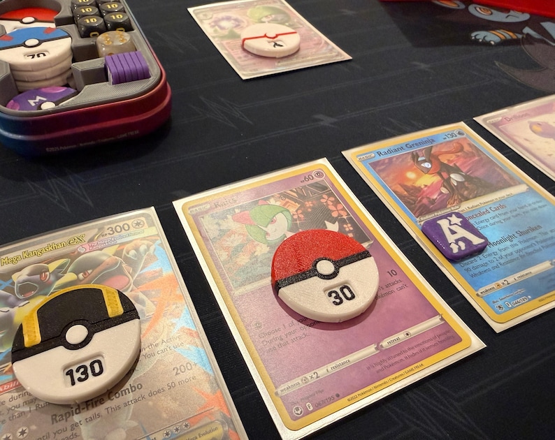 Pode incluir: Um conjunto de acess&oacute;rios para jogos de cartas colecion&aacute;veis, incluindo tr&ecirc;s contadores de dano circulares de pl&aacute;stico com detalhes em vermelho, preto, branco e amarelo, exibindo os n&uacute;meros 30 e 130, um marcador quadrado roxo com a letra A e um estojo.