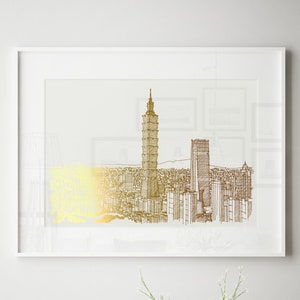 May include: A white framed print of a gold and brown line drawing of a city skyline with a tall building in the center. The print is hanging on a white wall above a wooden table with a vase of white flowers, candles, and a small concrete house figurine.