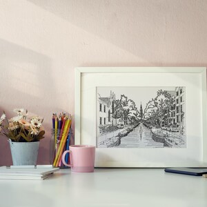 Puede incluir: Un dibujo enmarcado en blanco de una calle de la ciudad con un canal que la atraviesa. El dibujo est&aacute; en blanco y negro y est&aacute; rodeado de un paspart&uacute; blanco. El marco est&aacute; sobre un escritorio blanco con una taza rosa, un jarr&oacute;n de flores, una figurita de madera y una pila de libros.