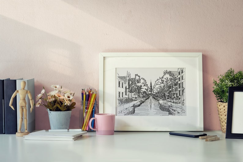 Puede incluir: Un dibujo enmarcado en blanco de una calle de la ciudad con un canal que la atraviesa. El dibujo est&aacute; en blanco y negro y est&aacute; rodeado de un paspart&uacute; blanco. El marco est&aacute; sobre un escritorio blanco con una taza rosa, un jarr&oacute;n de flores, una figurita de madera y una pila de libros.