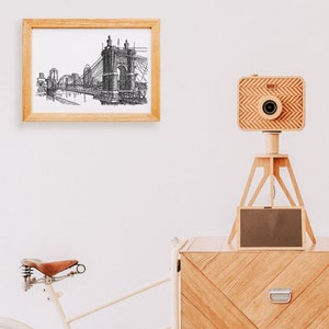 May include: A wooden camera on a tripod with a black and white drawing of a bridge in a wooden frame on a white wall. The camera is made of wood and has a lens and a shutter. The tripod is made of wood and has three legs. The drawing is of a bridge with a suspension bridge and a tower. The frame is made of wood and has a glass front.