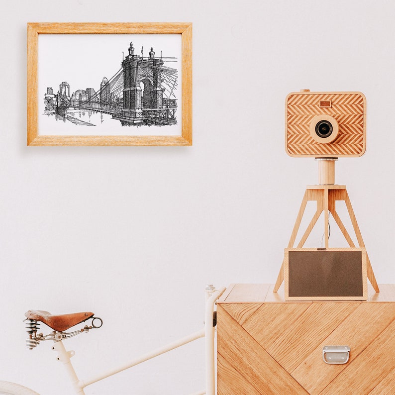 May include: A wooden camera on a tripod with a black and white drawing of a bridge in a wooden frame on a white wall. The camera is made of wood and has a lens and a shutter. The tripod is made of wood and has three legs. The drawing is of a bridge with a suspension bridge and a tower. The frame is made of wood and has a glass front.