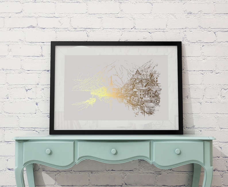 May include: Framed art print featuring a detailed illustration of a village nestled against a mountain range. The artwork uses gold and gray tones, with a focus on the architectural details of the buildings. The print is displayed in a black frame.