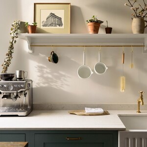 May include: A kitchen scene featuring a stainless steel espresso machine, white ceramic ladles, and a brass faucet. A framed architectural print and potted plants adorn a shelf above the countertop. A wooden cutting board and a white cloth are on the countertop.