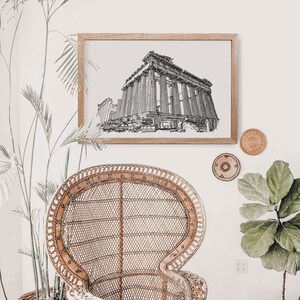 May include: A framed black and white architectural sketch of a classical building with columns. The artwork is in a light wood frame, hanging on a white wall. A woven wicker chair and decorative wall hangings are also visible.