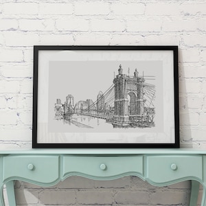 May include: Black and white line drawing of a bridge with buildings in the background, framed in a black frame and hanging on a white brick wall above a light blue dresser.
