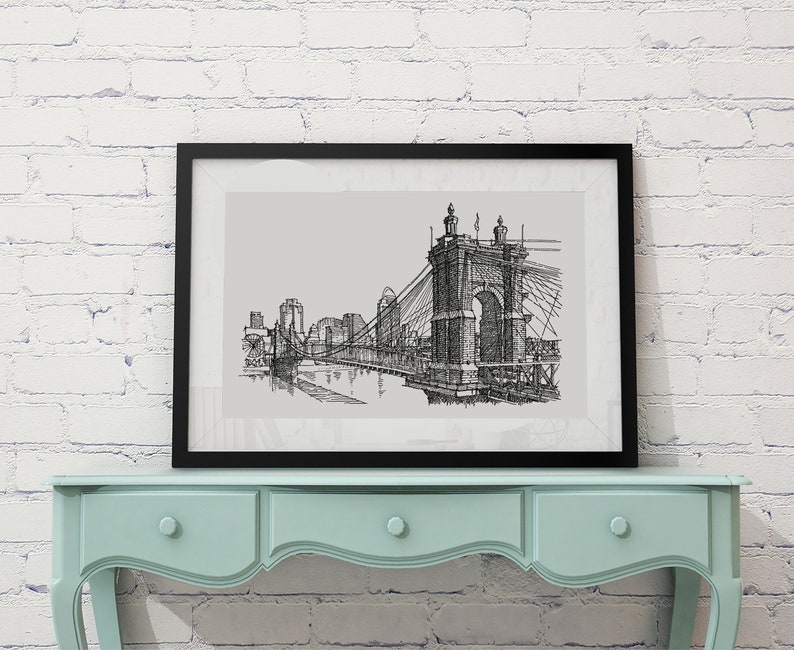 May include: Black and white line drawing of a bridge with buildings in the background, framed in a black frame and hanging on a white brick wall above a light blue dresser.