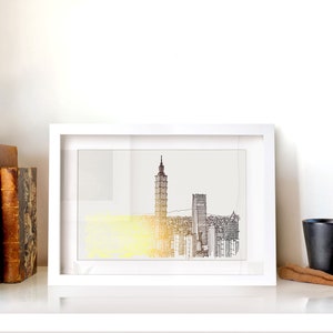 May include: A white framed print of a black and white line drawing of a city skyline with a tall building in the center. The print is on a white shelf with a small potted plant, books, and a black coffee mug.