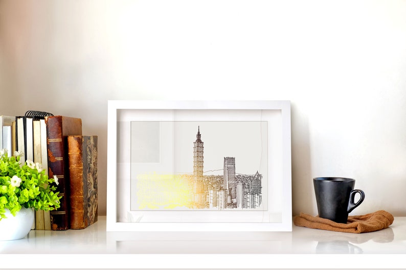 May include: A white framed print of a black and white line drawing of a city skyline with a tall building in the center. The print is on a white shelf with a small potted plant, books, and a black coffee mug.