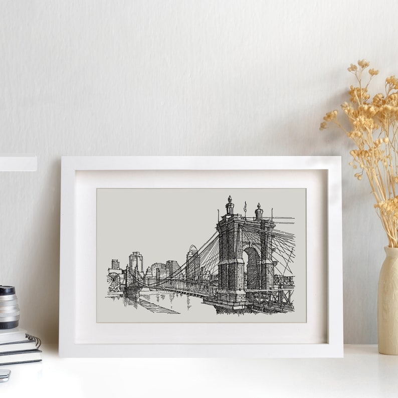 May include: Black and white line drawing of a suspension bridge with a cityscape in the background. The bridge is the focal point of the image, with the cityscape appearing in the distance. The drawing is framed in a white frame.