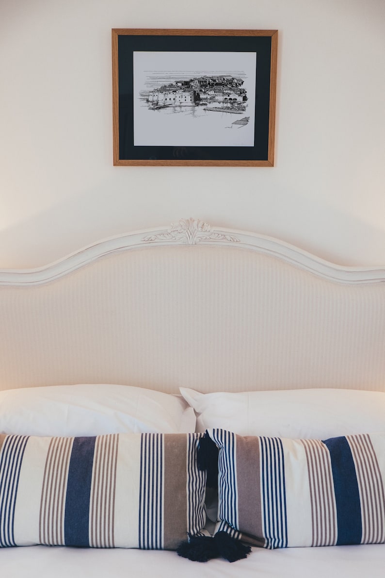 Puede incluir: Una cabecera tapizada en blanco con la parte superior curvada y un dibujo en blanco y negro enmarcado de una ciudad encima. La cama est&aacute; hecha con s&aacute;banas blancas y dos almohadas a rayas con borlas negras.