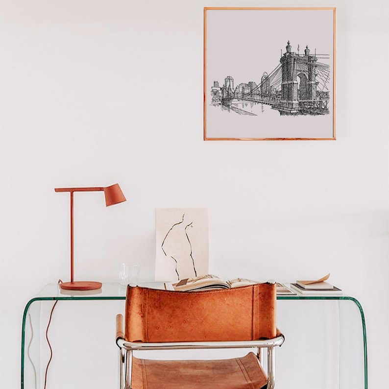 May include: A black and white line drawing of a bridge and cityscape hangs in a white room above a glass desk with a brown leather chair and a red lamp.