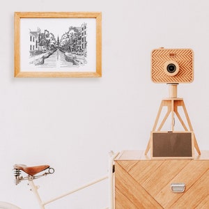 Puede incluir: Una c&aacute;mara de madera en un tr&iacute;pode con una pantalla negra, un dibujo en blanco y negro enmarcado de un canal y una c&oacute;moda de madera con un caj&oacute;n.