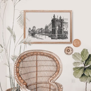 May include: Black and white line art print of a bridge with a cityscape in the background. The print is framed in a light wood frame and hanging on a white wall.