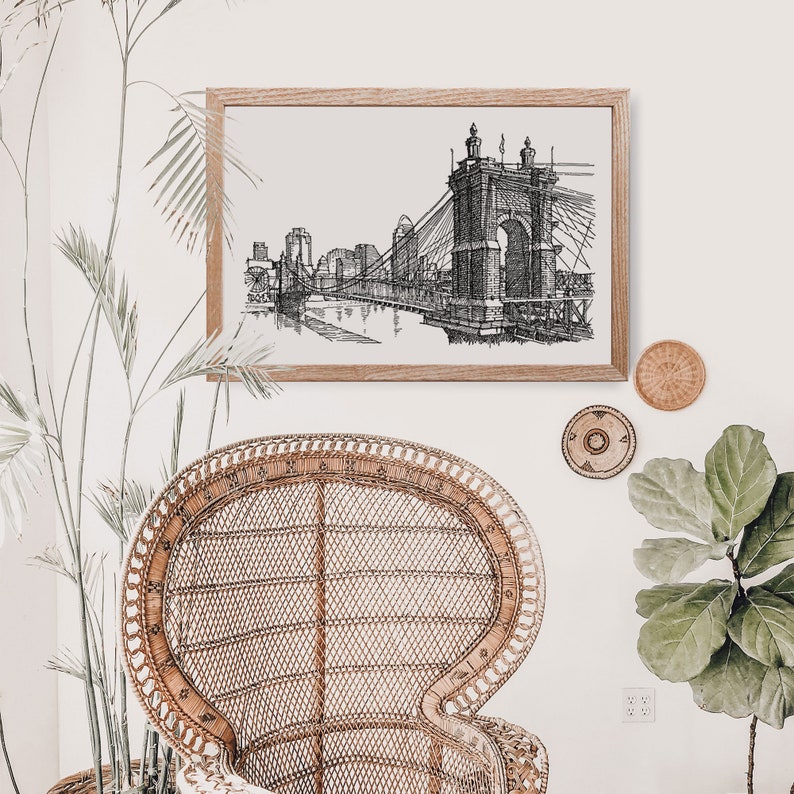 May include: Black and white line art print of a bridge with a cityscape in the background. The print is framed in a light wood frame and hanging on a white wall.