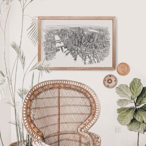May include: A black and white line drawing of a city skyline in a wooden frame hanging on a white wall. A wicker chair sits in front of the artwork.
