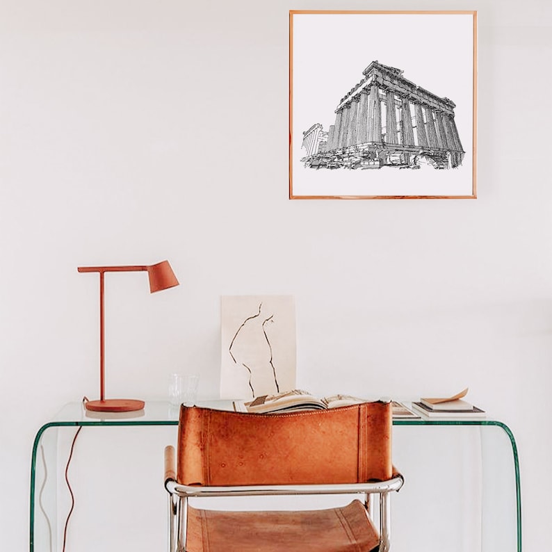 May include: A framed black and white architectural print of a classical building hangs on a white wall. A red desk lamp and a leather chair are in front of a glass desk. A drawing and an open book are on the desk.