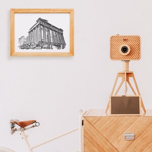 May include: A framed black and white sketch of a classical building hangs on a white wall. A wooden camera with a chevron pattern sits on a wooden tripod. A bicycle with a brown leather seat is partially visible.