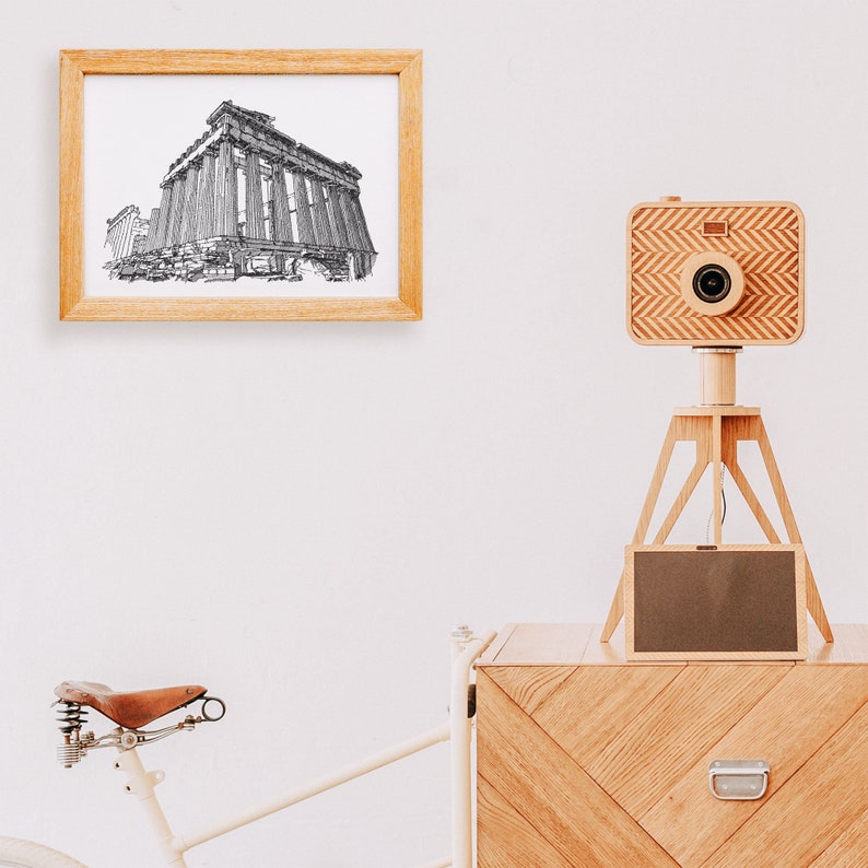 May include: A framed black and white sketch of a classical building hangs on a white wall. A wooden camera with a chevron pattern sits on a wooden tripod. A bicycle with a brown leather seat is partially visible.